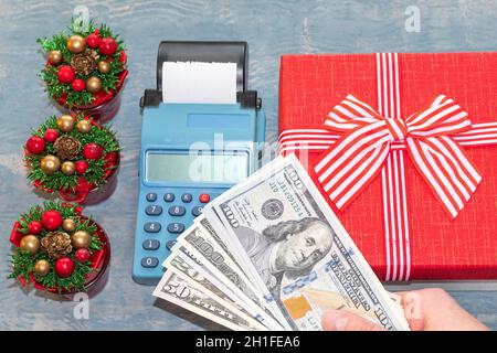Christmas sale. A hand holding dollars over a cash register near a red gift box with a ribbon and small Christmas trees with cones. online shopping. B Stock Photo