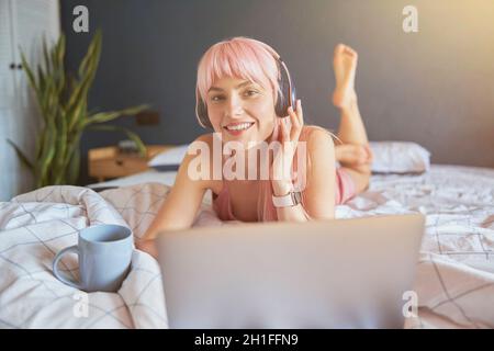 Cheerful woman with headphones and cup rests using laptop on comfortable bed Stock Photo