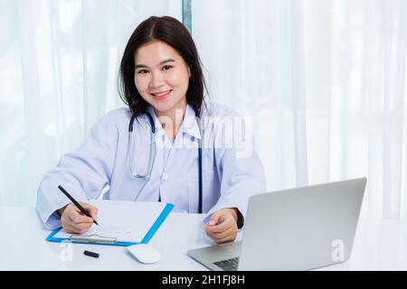 Asian doctor young beautiful woman smiling using working with a laptop computer and her writing something on paperwork or clipboard white paper at hos Stock Photo