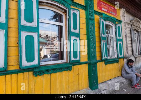 Young man sitting by a green and yellow painted traditional wooden house in use as restaurant in Ulan-Ude. Republic of Buryatia, Russia Stock Photo