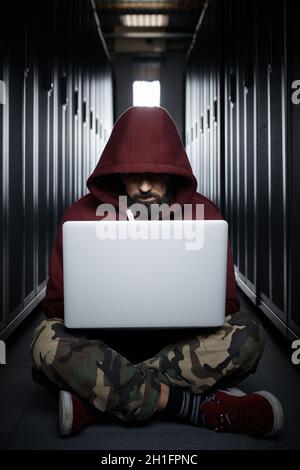 The hacker in a hood sits with the computer among servers, the burglar, face covered with a hood. Storage of data, cloudy services, system breaking Stock Photo