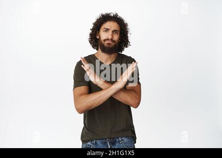 Disappointed young man making stop gesture, showing crossed arms sign, decline, prohibit and disagree, standing over white background Stock Photo