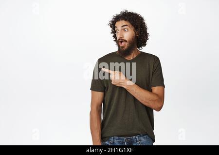 Portrait of surprised male model pointing and looking left, checking out big discount, showing sale, standing over white background Stock Photo