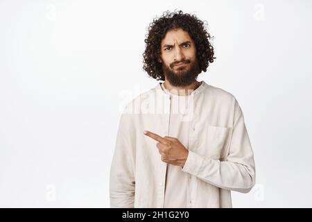 Sad and disappointed middle eastern guy pointing left, showing advertisement with upset face expression, white background Stock Photo