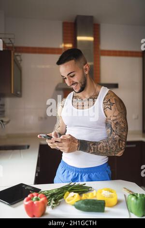 Vertical shot of a young tattooed man smiling at his phone in the kitchen Stock Photo