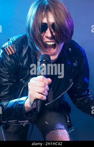 The young guy in black leather jacket is singing in microphone rock music on blue background. Stock Photo