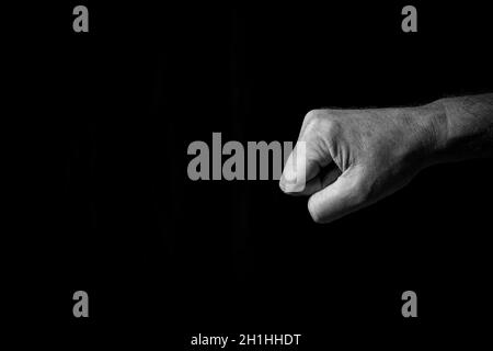 Black & white image of wrinkled male hand held out with clenched fist for fist bump or punch, isolated against a black background with dramatic lighti Stock Photo