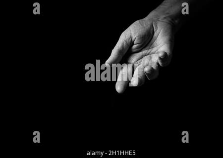 Black & white image of wrinkled male hand held out gesturing or offering help, isolated against a black background with dramatic lighting. Stock Photo