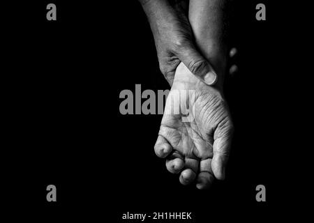 Black & white image of wrinkled male hands holding wrist with open palm, isolated against a black background with dramatic lighting. Stock Photo