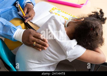 Education Preschool 3-4 year olds girl laying head on table, sad or tired at start of school day, teacher patting her on the back, wearing face mask Stock Photo