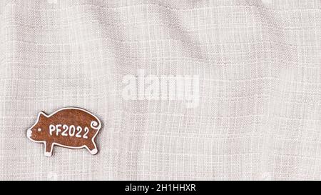 Sweet gingerbread piggy for 2022 happy New Year on a creased textile background. Xmas cookie in pig shape on decorative texture of wavy beige fabric. Stock Photo