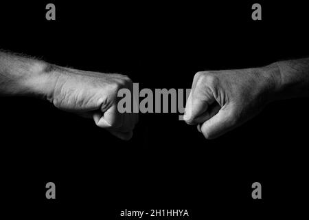 Black & white image of wrinkled male hands held out with clenched fists for fist bump or punch, isolated against a black background with dramatic ligh Stock Photo