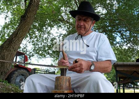 Farmer with hammer and iron tool on the tree stump is sharpening his scythe in Trnovec, Croatia Stock Photo