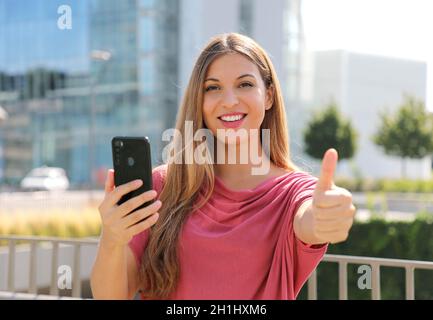 Happy beautiful woman thumbs up with smartphone in her hand and modern city on background. Positive businesswoman smiles at camera. Stock Photo