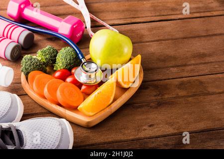 Top view of fresh fruits and vegetables in heart plate wood (apple, carrot, tomato, orange, broccoli) and sports equipment and doctor stethoscope on w Stock Photo