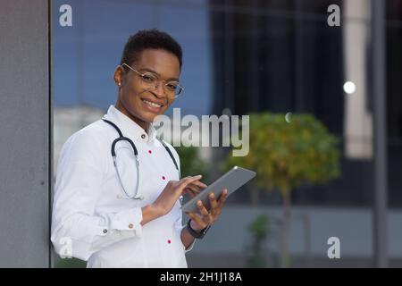 Portrait of young beautiful african american doctor woman smiling happy looking at camera and holding tablet, for patient consultation online near cli Stock Photo