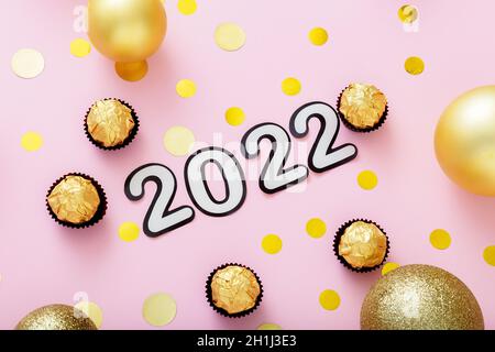 2022 numeral lettering in Happy New Year style. 2022 text with Christmas decorations candies Sweets gold confetti on pink color background. Stock Photo