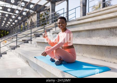 African American woman smiling and looking at camera, sitting in lotus position on fitness mat, confident and cheerful after yoga Stock Photo