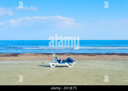 LARNACA, CYPRUS - FEB 18, 2019: Woman taking sunbathes on a deserted beach. Cyprus is a famous tourist destination. Stock Photo