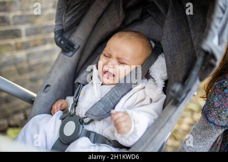 Picture of a distressed baby in gray clothing crying with eyes closed in a dark gray stroller on a sidewalk covered by leaves and next to a brick wall Stock Photo