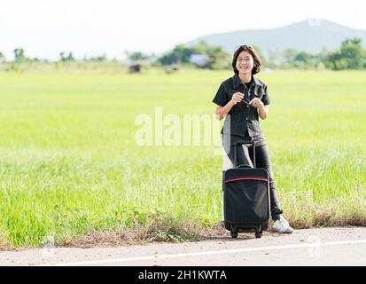 Young asian woman short hair and wearing sunglasses with luggage hitchhiking along a road in countryside Thailand Stock Photo