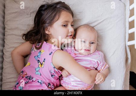 Happy little girl lovingly hugging baby sister in pink Asian attire on a bed. Two lovely siblings in pink attires lying down on bed. Young siblings ha Stock Photo