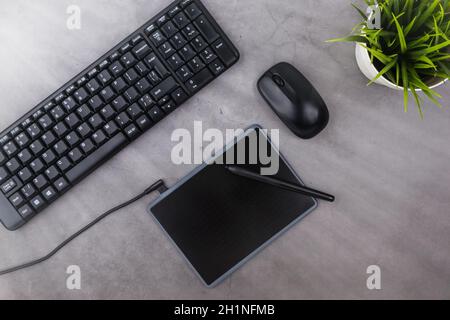 Working place on dark table with copy space. Keyboard, computer mouse, pen, graphics tablet, flower in pot. Flat lay. Top view, the sun's rays from th Stock Photo