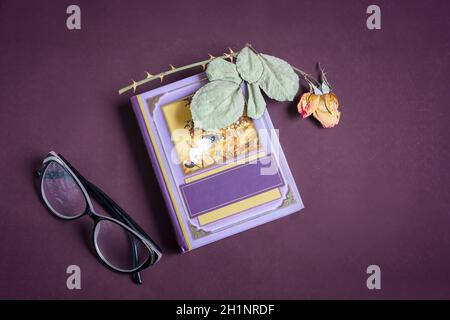 On a small book is a dried rose flower with leaves. There are glasses next to it. Presented on a dark background. Top view with space to copy, flat la Stock Photo