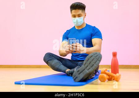 Young Latin man with protective face mask, relaxed, using mobile phone after workout in gym during pandemic new normal. High quality photo Stock Photo