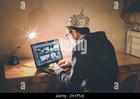 Young man with aluminum cap is sitting in the dark basement in front of a laptop. Conspiracy theory concept Stock Photo