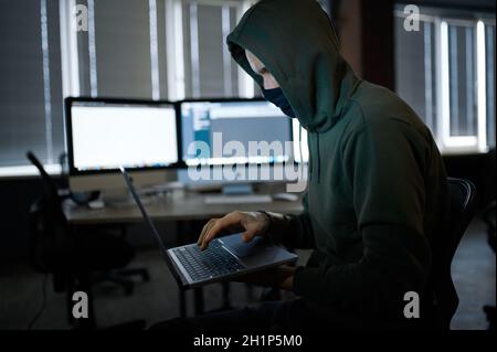 Male internet hacker in hood holds laptop, front view. Illegal web programmer at workplace, criminal occupation. Data hacking, cyber safety Stock Photo