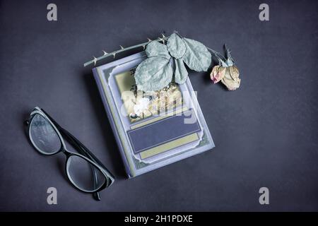 On a small book is a dried rose flower with leaves. There are glasses next to it. Presented on a dark background. Top view with space to copy, flat la Stock Photo