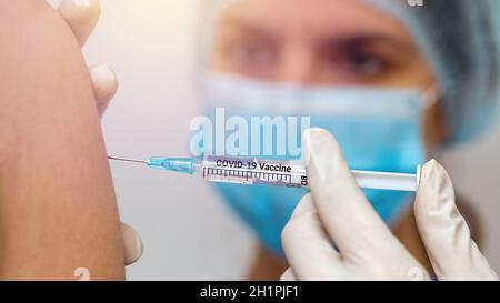 Female nurse in blue surgical mask injecting vaccine against covid-19 into patient's shoulder from close-up. Concept of fighting coronavirus pandemic. Stock Photo