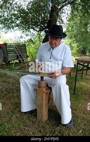 Farmer with hammer and iron tool on the tree stump is sharpening his scythe in Trnovec, Croatia Stock Photo