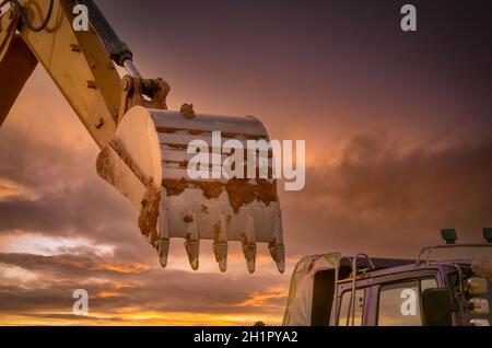 Dirt metal bucket of backhoe after digging soil. Backhoe parked near truck with golden sunset sky background. Crawler excavator. Earthmoving machine a Stock Photo