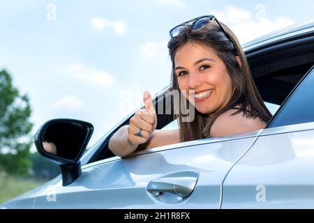 Close up portrait of young female car driver doing thumbs up in car. Woman looking backwards out of window. Stock Photo