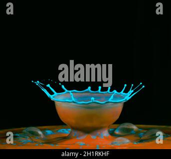 A blue water drop falls into a pool of colored water and breaks the surface as a bowl shape - Liquid Drop Art Stock Photo