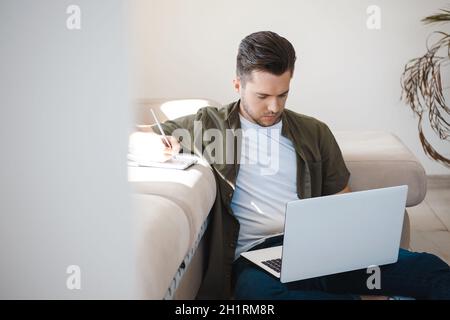 Man participating an online conference sitting on the floor and taking notes in his notebook. Looking down Stock Photo