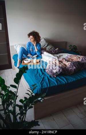 Caucasian woman sitting in blue pajamas in bed looking back and falling to thoughts while a healthy breakfast awaits her on the bed. Breakfast in bed Stock Photo