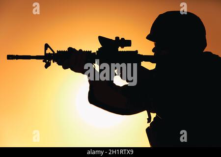 Silhouette of modern army infantry soldier standing on background of setting sun and aiming assault rifle with collimator sight. Special operations ri Stock Photo