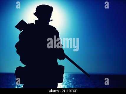 Silhouette of army elite forces fighter standing with sniper rifle on background of blue sky with moon or sun. Sniper or marksman in boonie hat, carry Stock Photo