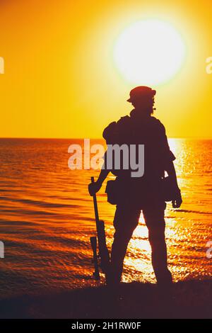 Silhouette of army commando fighter, Navy SEALS team sniper or coast guard soldier standing on sea shore with rifle on background of ocean sunset hori Stock Photo