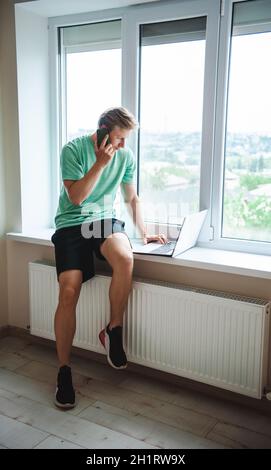 Side view of a man sitting on the windowsill talking on the phone with his co-worker while looking for plane tickets for a vacation at sea on his