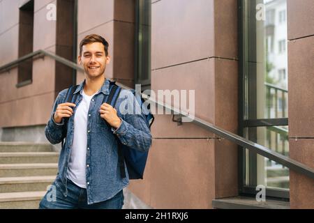 Medium shot portrait of cheerful handsome young male of food delivery service with large thermo backpack standing posing in office building stairs Stock Photo