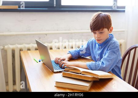 Teen boy doing homework online at home. He looks at the textbook and types a message with questions to the teacher. Online education concept. Stock Photo