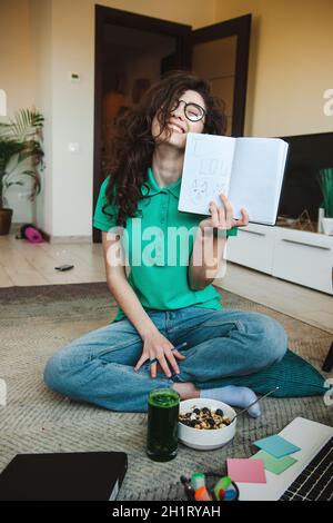 Women having fun in her room sitting on the floor and holding an open notebook in her left hand pointing to the camera that she loves cats. Smiling Stock Photo