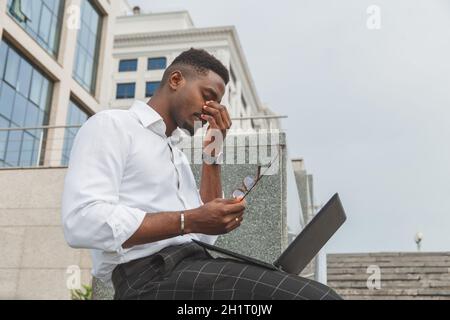 Feeling exhausted. Frustrated young black african man keeping eyes closed and looking tired from extra working on laptop Stock Photo