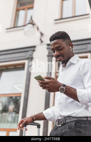 Young handsome black man with luggage using smartphone and listening music with earphones in the city. Stock Photo