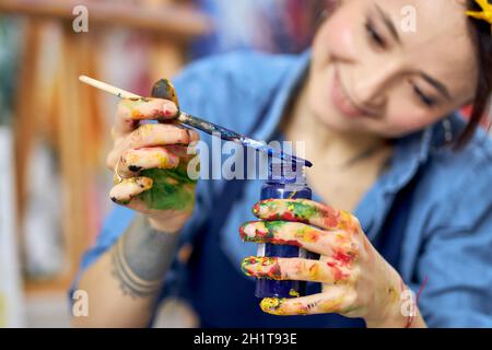 Smiling young woman in apron holding blue paint jar and a paintbrush, working on a painting in workshop Stock Photo