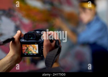 Close up of male hands holding professional digital camera on a blurred background. Female artist creating painting in the background Stock Photo